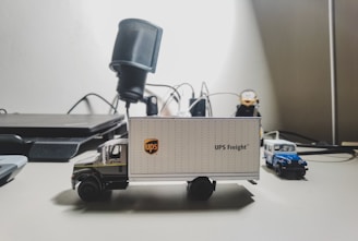 A detailed miniature model of a UPS delivery truck sits on a desk, with various office items in the background. The truck features the UPS logo and is accompanied by a smaller blue and white toy vehicle.