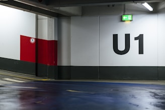 An indoor parking lot with a large U1 sign painted on the wall. The environment features concrete walls with a grey base and a red and white section. A green exit sign with an arrow points left, and the floor is coated with a blue finish. There is a pedestrian control sign on the red section of the wall.