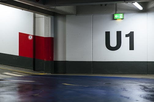 An indoor parking lot with a large U1 sign painted on the wall. The environment features concrete walls with a grey base and a red and white section. A green exit sign with an arrow points left, and the floor is coated with a blue finish. There is a pedestrian control sign on the red section of the wall.
