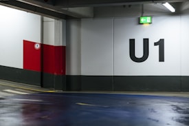 An indoor parking lot with a large U1 sign painted on the wall. The environment features concrete walls with a grey base and a red and white section. A green exit sign with an arrow points left, and the floor is coated with a blue finish. There is a pedestrian control sign on the red section of the wall.