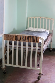An old-fashioned hospital bed with wheels is placed in a sparsely decorated room. A cardboard sign reading 'Contagious Disease Quarantine' is attached to the foot of the bed. The bed is made with a simple white pillow and a grey blanket. The room has plain light green walls and a tiled floor.