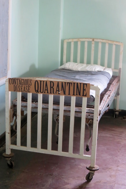 An old-fashioned hospital bed with wheels is placed in a sparsely decorated room. A cardboard sign reading 'Contagious Disease Quarantine' is attached to the foot of the bed. The bed is made with a simple white pillow and a grey blanket. The room has plain light green walls and a tiled floor.