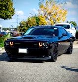 A sleek black muscle car with distinctive circular headlights is parked on a road. Surrounding the car are autumn trees with vibrant yellow and green foliage, against a backdrop of a bright blue sky. Another vehicle is partially visible behind it.