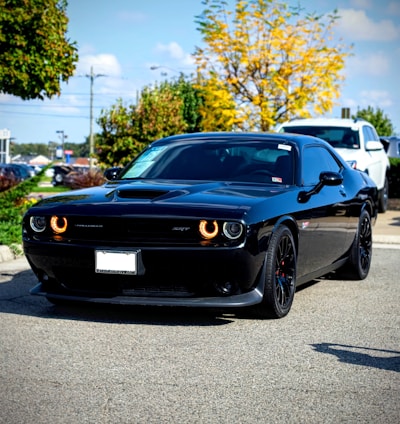 A sleek black muscle car with distinctive circular headlights is parked on a road. Surrounding the car are autumn trees with vibrant yellow and green foliage, against a backdrop of a bright blue sky. Another vehicle is partially visible behind it.