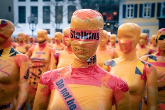 A crowd of mannequin figures wrapped in orange and red tape, with inscriptions like 'Stalking' and 'Do not be silent'. These figures are placed closely together outdoors in front of buildings, creating an impactful visual statement.