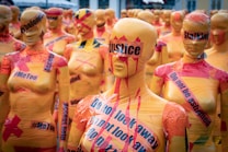 An assembly of mannequins covered in yellow and red tape, each bearing various messages related to social justice issues. The tape features words like 'justice,' '#MeToo,' 'Do not look away,' and 'Stalking.' The mannequins stand closely together, conveying a strong visual impact.