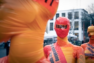 Mannequins covered in orange and yellow tape with slogans are displayed in an outdoor urban area. Background shows a few buildings and bare trees.