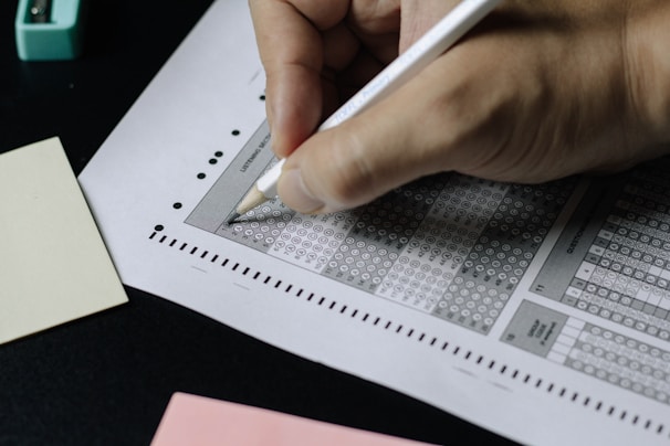 A hand holding a pencil is filling out a multiple-choice answer sheet, typically used for standardized tests. The sheet features a grid of circles labeled with letters, and some circles are filled in. Nearby, there are pink and yellow index cards.