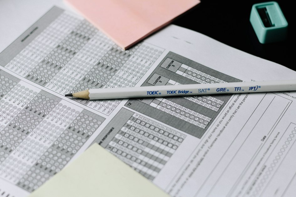 A pencil resting across a printed multiple-choice answer sheet covered in rows of bubbles, with a second form and sticky notes underneath