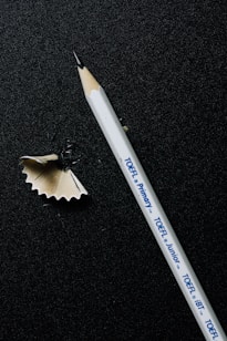 A sharpened white pencil lies on a dark textured surface, alongside a curled wood shaving. The pencil has printed text indicating different types of TOEFL exams.