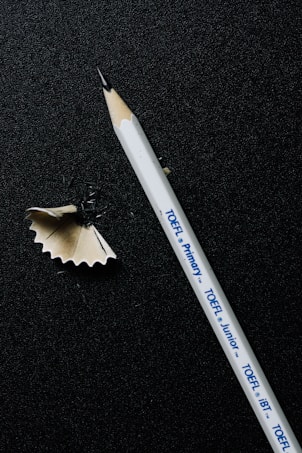 A sharpened white pencil lies on a dark textured surface, alongside a curled wood shaving. The pencil has printed text indicating different types of TOEFL exams.