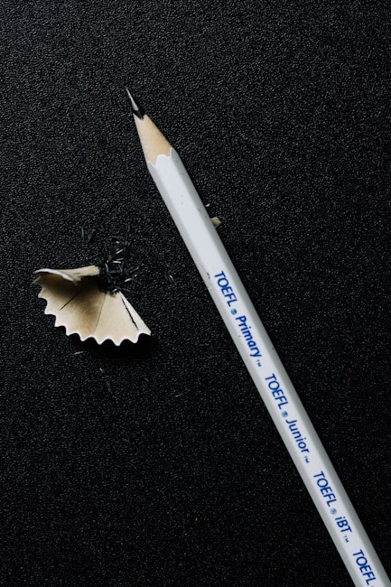 A sharpened white pencil lies on a dark textured surface, alongside a curled wood shaving. The pencil has printed text indicating different types of TOEFL exams.