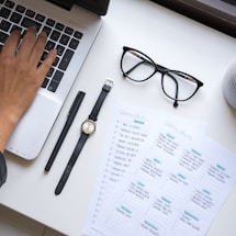 A hand is typing on a laptop keyboard placed on a white desk. Nearby, a pair of black-framed glasses, a black pen, a wristwatch with a black strap, and two sheets of paper with handwritten notes can be seen. The sheets include a personal or work to-do list.