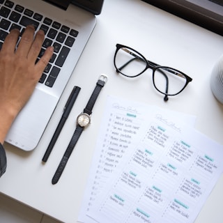 A hand is typing on a laptop keyboard placed on a white desk. Nearby, a pair of black-framed glasses, a black pen, a wristwatch with a black strap, and two sheets of paper with handwritten notes can be seen. The sheets include a personal or work to-do list.