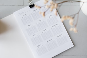 An open planner page displaying a calendar layout for the entire year, written in Spanish. Months are arranged in a grid format. A black binder clip is attached to the top of the page. A branch with delicate, dry flowers is blurred in the foreground, adding a soft touch. The background is neutral and minimalistic.
