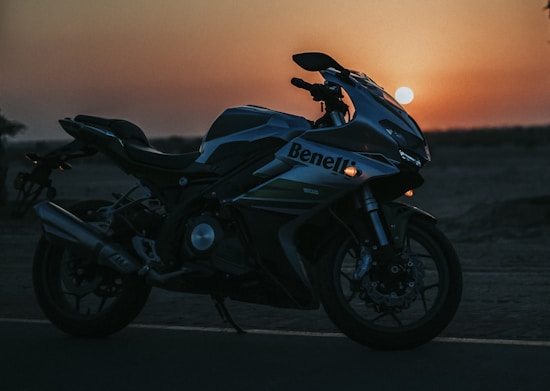 A motorcycle is parked on a road during sunset, highlighted against the vibrant orange and red hues of the sky. The silhouette of the bike is detailed, showcasing its sleek design and sporty features.