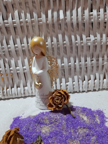 A small angel figurine with golden wings and a heart pendant stands against a wicker background. The figurine is surrounded by dried brown roses and scattered purple sand. The setting is soft and serene, suggesting a decorative or nostalgic theme.