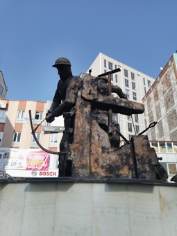 A metal sculpture of a worker wearing a helmet, operating a large industrial machine or press. The sculpture is positioned in an urban setting with buildings in the background under a clear blue sky. Advertisements and signs are visible on the buildings behind.
