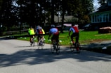 Four cyclists wearing bright jerseys ride along a quiet road surrounded by greenery and houses. The scene shows a leisurely activity in a suburban setting with trees casting shadows on the pavement.