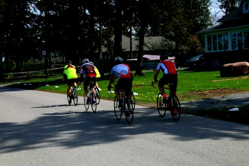 Four cyclists wearing bright jerseys ride along a quiet road surrounded by greenery and houses. The scene shows a leisurely activity in a suburban setting with trees casting shadows on the pavement.