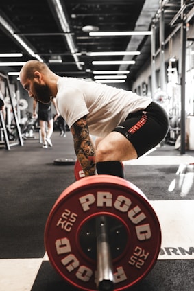 man in white t-shirt and black shorts carrying red and black barbell