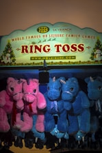 A vibrant carnival game sign for 'Ring Toss' is prominently displayed. It features festive decorations, including Christmas themes with a snowman and a Christmas tree. Below the sign, several plush unicorn toys are hanging as prizes, with vivid pink and blue colors.