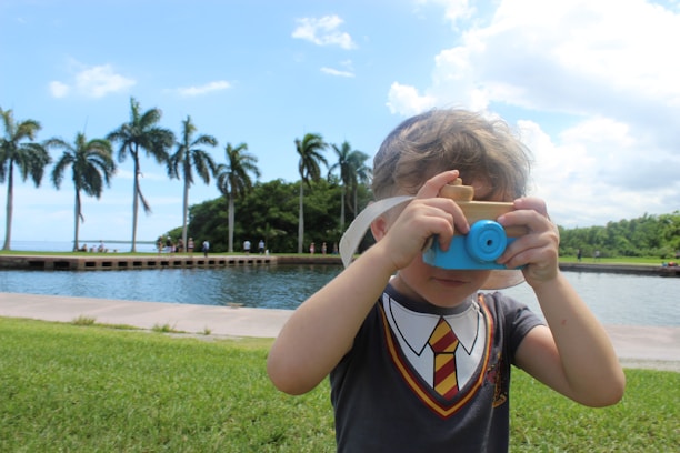 A young child wearing a shirt with a tie design is holding a small toy camera to their face. They are standing on a grassy area beside a body of water with a row of palm trees and a bright blue sky in the background.