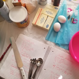 A table is set with various baking supplies and tools, including sticks of unsalted butter, a container of shortening, two eggs, a can of baking powder, and a collection of measuring spoons. There are also some piping bags, decorating tips, a pink mixing bowl, and a spatula. In the center, a notebook with handwritten recipes is open, each recipe written in red ink.