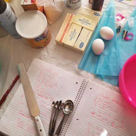 A table is set with various baking supplies and tools, including sticks of unsalted butter, a container of shortening, two eggs, a can of baking powder, and a collection of measuring spoons. There are also some piping bags, decorating tips, a pink mixing bowl, and a spatula. In the center, a notebook with handwritten recipes is open, each recipe written in red ink.