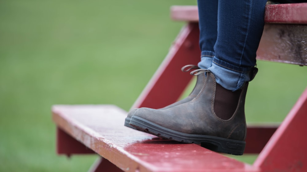 person wearing blue denim jeans and black leather boots sitting on red wooden bench