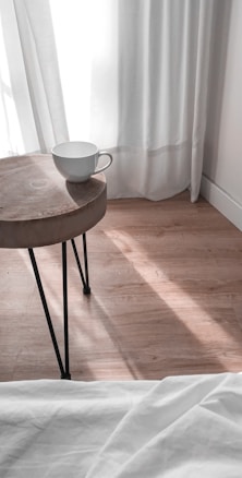 A simple, minimalist scene featuring a wooden round table with black metal legs, on which rests a white mug. The background displays white curtains with sunlight streaming through, casting soft shadows on a light wooden floor. The edge of a white bedspread is visible at the bottom of the image.