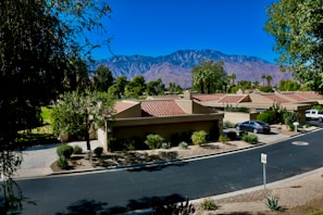 A suburban neighborhood with a row of tan houses featuring red-tiled roofs sits in a lush green setting. A paved road curves gently through the scene, bordered by trimmed bushes and trees. In the background, majestic mountains rise under a clear blue sky. Parks and green spaces surround the homes, adding to the tranquil environment. A few vehicles are parked in driveways, and a speed limit sign is visible on the road.
