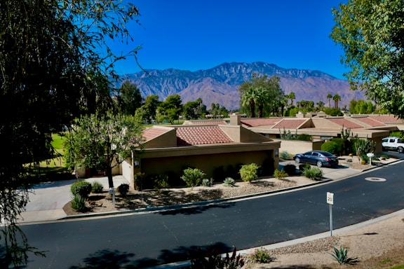 A suburban neighborhood with a row of tan houses featuring red-tiled roofs sits in a lush green setting. A paved road curves gently through the scene, bordered by trimmed bushes and trees. In the background, majestic mountains rise under a clear blue sky. Parks and green spaces surround the homes, adding to the tranquil environment. A few vehicles are parked in driveways, and a speed limit sign is visible on the road.