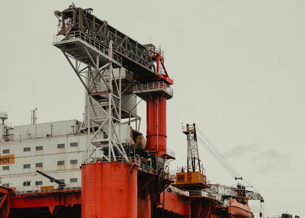 An industrial offshore oil platform featuring large, red structural components and intricate metal frameworks. The platform includes cranes, staircases, and multiple levels, with a beige-painted building section displaying numerous windows.
