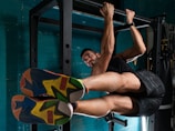 A man is performing an exercise on a pull-up bar, engaging his core muscles with his legs raised horizontally. He is wearing brightly colored athletic shoes and sportswear, and the setting appears to be a gym with teal walls.