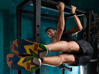 A man is performing an exercise on a pull-up bar, engaging his core muscles with his legs raised horizontally. He is wearing brightly colored athletic shoes and sportswear, and the setting appears to be a gym with teal walls.