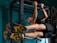 A man is performing an exercise on a pull-up bar, engaging his core muscles with his legs raised horizontally. He is wearing brightly colored athletic shoes and sportswear, and the setting appears to be a gym with teal walls.