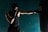 A person wearing boxing gloves and a sleeveless top is shown in a boxing stance, striking a heavy bag. The setting includes a textured teal wall in the background, casting shadows that create a dramatic effect.
