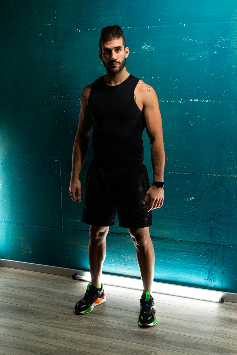 Hombre con camiseta sin mangas negra y pantalones cortos de pie junto a la pared azul