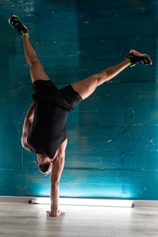A person is performing a handstand against a textured teal wall, with a visible focus on balance and strength. The athlete is wearing athletic shoes and dark sportswear, with a small horizontal light fixture on the floor beneath.