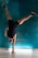 A person is performing a handstand against a textured teal wall, with a visible focus on balance and strength. The athlete is wearing athletic shoes and dark sportswear, with a small horizontal light fixture on the floor beneath.