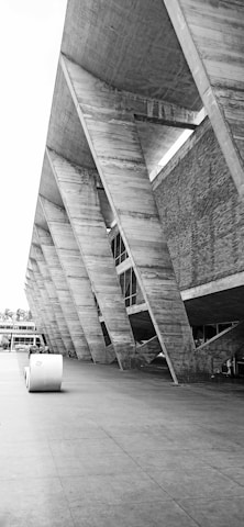 A striking piece of modern architecture with large concrete beams and a geometric design. The structure features a series of angular supports that create an imposing and futuristic appearance. The exterior wall combines concrete and brick elements, offering a textured look.