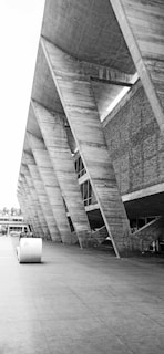 A striking piece of modern architecture with large concrete beams and a geometric design. The structure features a series of angular supports that create an imposing and futuristic appearance. The exterior wall combines concrete and brick elements, offering a textured look.