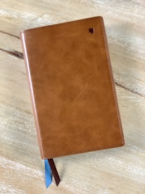 A closed leather-bound book with a textured brown cover, featuring a small logo in the top right corner. Two ribbon bookmarks, one blue and one brown, extend from the bottom. The book is resting on a light wooden surface.