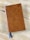 A closed leather-bound book with a textured brown cover, featuring a small logo in the top right corner. Two ribbon bookmarks, one blue and one brown, extend from the bottom. The book is resting on a light wooden surface.