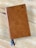 A closed leather-bound book with a textured brown cover, featuring a small logo in the top right corner. Two ribbon bookmarks, one blue and one brown, extend from the bottom. The book is resting on a light wooden surface.