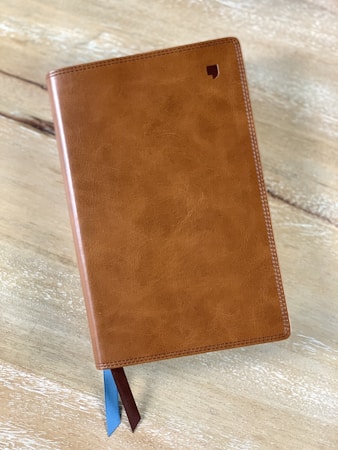 A closed leather-bound book with a textured brown cover, featuring a small logo in the top right corner. Two ribbon bookmarks, one blue and one brown, extend from the bottom. The book is resting on a light wooden surface.