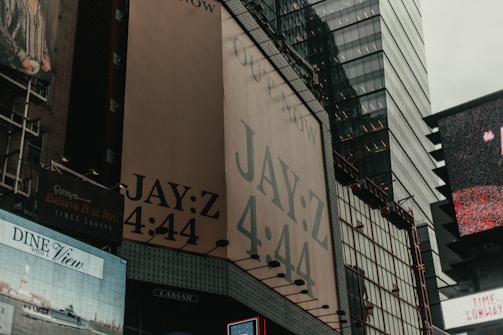 A cityscape view featuring large billboards on tall buildings, prominently displaying an advertisement for Jay-Z's '4:44' album. The scene includes various other signs and advertisements, with reflections visible on glass surfaces of surrounding skyscrapers.