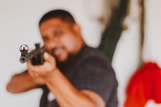 A person holds and points a rifle towards the camera with their face and upper body blurred. The focus is on the barrel of the gun and the person is wearing dark clothing. The background includes some greenery and a bright red fabric.