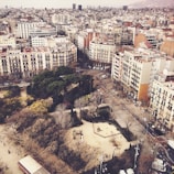An aerial view of an urban neighborhood featuring a mix of residential and commercial buildings. The streets are lined with trees, and a park with trees and pathways is visible in the foreground. The buildings are mostly multi-story, displaying a variety of architectural styles. The atmosphere appears calm and the city extends to the horizon.